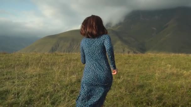 Una joven con un vestido azul corre colina abajo sobre el fondo de las montañas nubladas del Cáucaso. Movimiento lento. — Vídeos de Stock