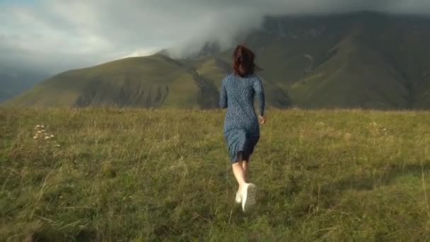 Una joven con un vestido azul corre colina abajo sobre el fondo de las montañas nubladas del Cáucaso. Movimiento lento. — Vídeo de stock