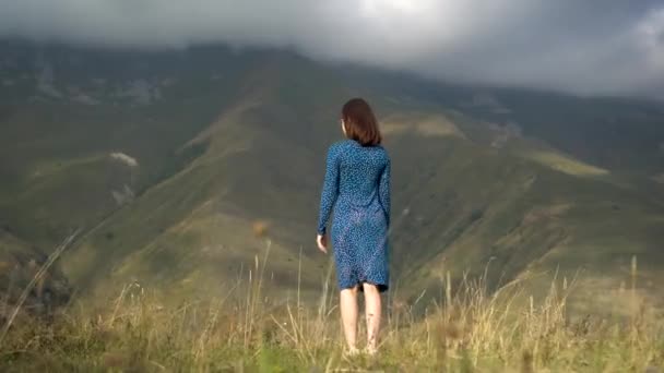 Een jonge vrouw staat in een blauwe jurk en heft haar handen omhoog kijkend naar de bergen. — Stockvideo