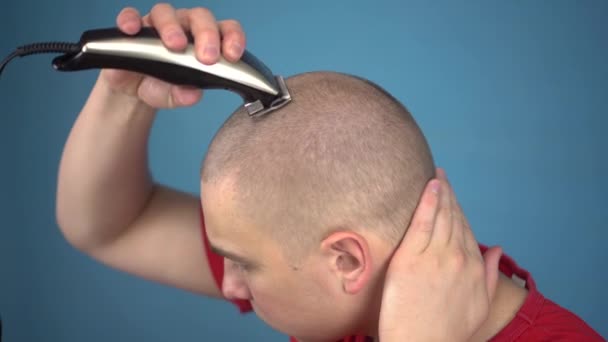 Kel genç adam küstahça başını tıraş eder. Bir adam saçını mavi arka planda bir kuaför makinesiyle kesiyor.. — Stok video