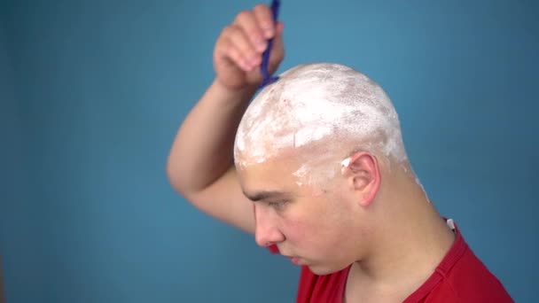 Den skalliga unge mannen rakar oförskämt sitt huvud. En man klipper sitt hår med en rakkniv på en blå bakgrund. — Stockvideo