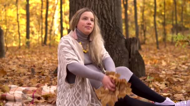 Eine schwangere Frau sitzt unter einem Baum und wirft umgefallenes Laub in den Herbstwald. Zeitlupe — Stockvideo