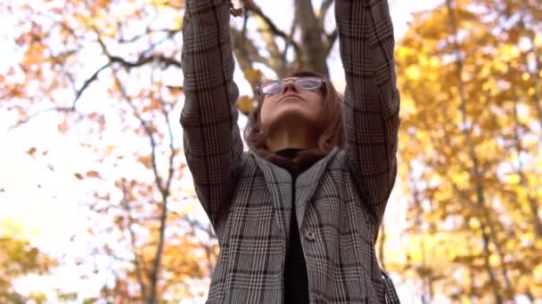 Een jonge vrouw met een bril kotst gele bladeren op in het herfstbos. Langzame beweging — Stockvideo