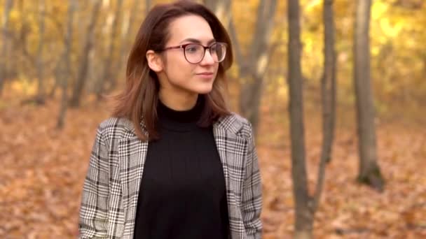 Egy szemüveges, kabátos fiatal nő sétál az őszi erdőben. Sárga levelek vannak itt. Lassú mozgás. — Stock videók