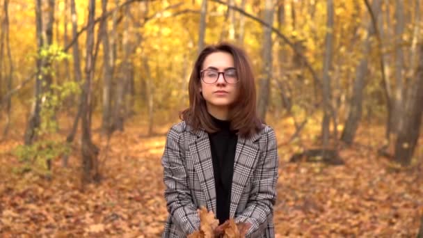 Молодая женщина в очках бросает желтые листья в осенний лес. Медленное движение — стоковое видео