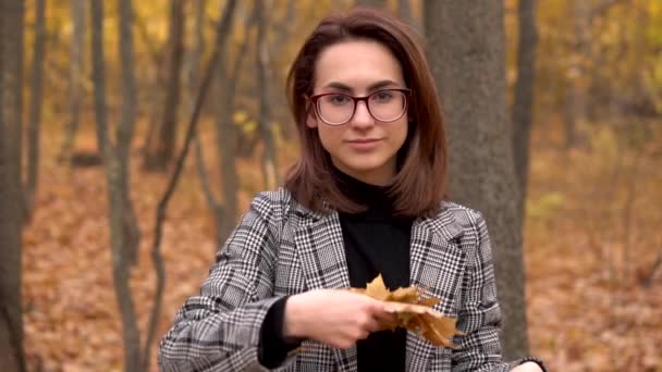 Молодая женщина в очках бросает желтые листья в осенний лес в камеру. Медленное движение — стоковое видео