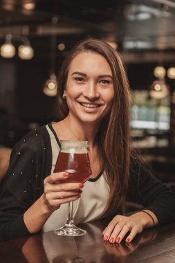 Vertical portrait of a happy beautiful woman toasting with her beer glass, smiling joyfully. Gorgeous young long haired woman cheering with a glass of craft beer. Brewery, consumerism concept clipart