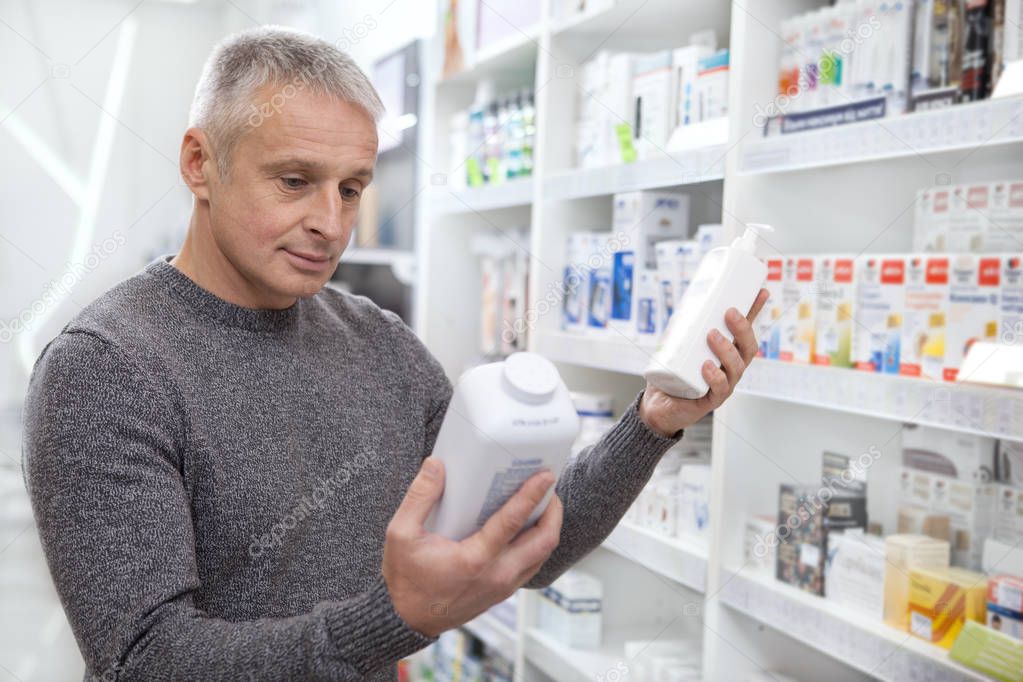 Senior male customer choosing between two medical products, shopping for goods at the drugstore. Mature man examining labels of two bottles of medications. Lifestyle, health, purchasing concept