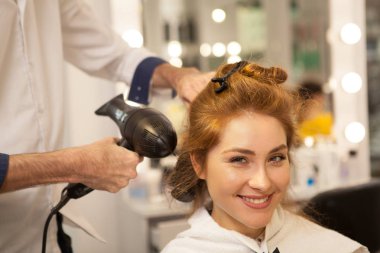 Kameraya heyecanla, uzun saçlarını styling Kuaför gülümseyen mutlu genç güzel kadın. Profesyonel kuaför kadın müşteri, kopya alanı ile çalışma. Kurutma, darbe saç modeli kavramı