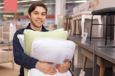 Neşeli genç adam kameraya gülümseyen, yumuşak yeni yastık yığını tutan Ev mobilya mağazasında satın alıyor. Eleketrik Yatak takımlar Bölümü'nde alışveriş çekici erkek müşteri yeni yastıklar, grup satın alma