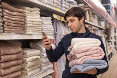 Tekstil bölümü havlulara için alışveriş yaparken onun akıllı telefon kullanan yakışıklı genç adam. Erkek müşteri online tarama, cep telefonuyla, linens süpermarkette satın alma. Teknoloji kavramı