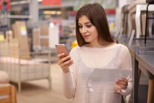 Attractive young woman using her smart phone at furniture department, while shopping for home goods. Female customer browsing online, checking price of an item using her phone at furnishings store