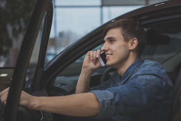 Young handsome man speaking on his phone, sitting in a new automobile at the dealership. Attractive cheerful man calling using his phone, after buying new car. Technology, mobility concept