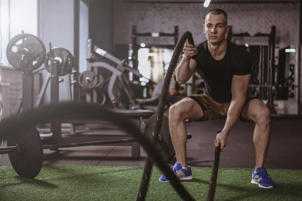 Young fitness man working out at crossfit box, exercising with battle ropes. Handsome crossfit athlete training at functional fitness gym, copy space. Sportsman doing functional battle rope workout