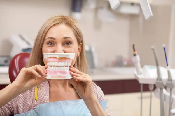 Happy mature female patient smiling posing with a big dentures mold in front of her mouth. Cheerful woman having fun at the dentists office, waiting for medical checkup by her dentist. Teeth care