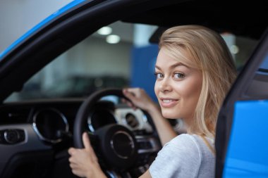 Attractive blond haired female driver smiling over her shoulder to the camera with her hands on the steering wheel of a new automobile. Happy woman sitting comfortably in a new car. Driving vehicle clipart