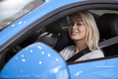 Beautiful female sitting in her new car behind the steering wheel, smiling to the camera confidently. Gorgeous woman driving blue car. Happy female enjoying comfortable automobile. Travel, safety clipart