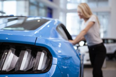 Selective focus on car lights on the foreground, woman looking inside the vehicle on the background, copy space. Female customer examining a car on sale at the dealership. Consumerism, driving concept clipart
