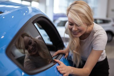 Long haired beautiful woman examining modern car on sale at the dealership showroom. Happy female customer checking out blue automobile at the car salon. Driving, ownership, buying cars concept clipart