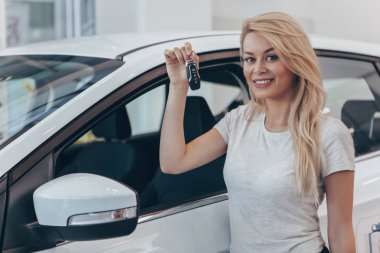 Young happy female driver holding car key, posing near her new automobile at dealership salon, copy space. Charming woman smiling to the camera after buying new auto, copy space. Owner, driver concept clipart