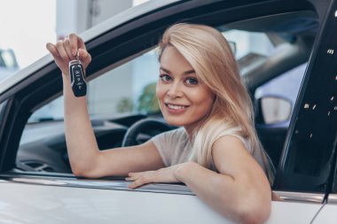 Happy young woman smiling joyfully to the camera, enjoying sitting in her new automobile at the dealership salon. Beautiful female driver holding car keys, sitting in a new car. Driving, safety concep clipart