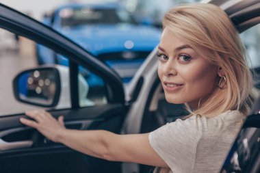 Onun omuz üzerinden kameraya gülümseyen, yeni bir Otomobil araba salon, kopya alanı oturan mutlu güzel kadın. Kadın seçerek yeni otomatik araba galerisinde çarpıcı. Kadınlık, güven