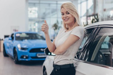 Excited young beautiful female driver posing near her newly bought automobile at the dealership, showing thumbs up, copy space. Gorgeous woman buying new car, smiling happily. Rental service, retail clipart