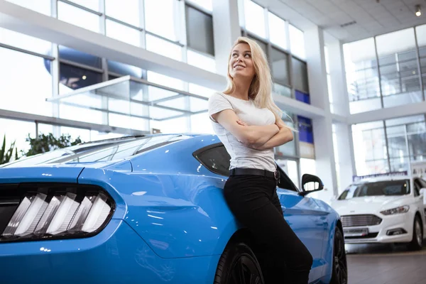 Low angle shot of stunning young female driver posing with her new automobile at the dealership salon. Happy woman looking away dreamily, leaning on a modern muscle car, copy space. Buying car concept