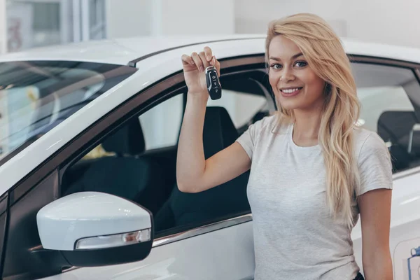 Young happy female driver holding car key, posing near her new automobile at dealership salon, copy space. Charming woman smiling to the camera after buying new auto, copy space. Owner, driver concept