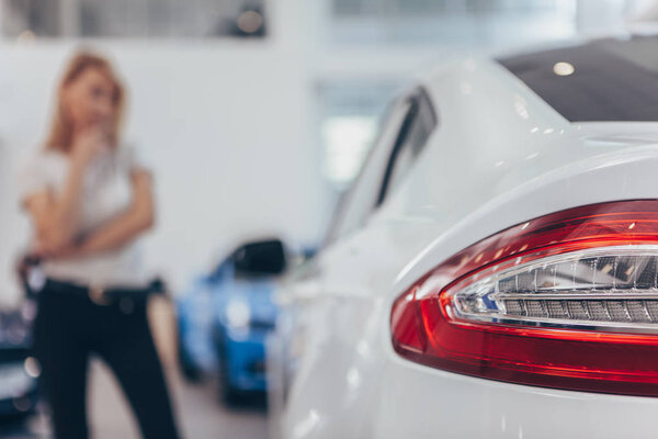 Selective focus on car lights of a modern automobile at the dealership, female customer looking at the auto on the background. Woman buying new car at dealership salon, copy space. Consumerism concept