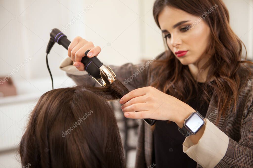 Beautiful female hairstylist working at her salon, curling hair of a female client