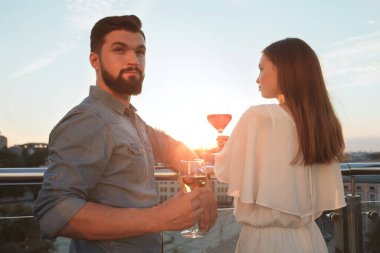 Yakışıklı sakallı adam gün batımında kız arkadaşıyla çatı katındaki restoranda şarap içiyor.