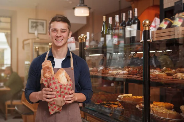 幸せなハンサムな若い男が笑って パン屋で働くことを楽しんで 新鮮なパンを販売する — ストック写真
