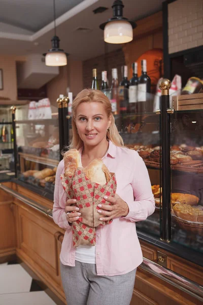 焼きたてのパンを持っている陽気な成熟した女性の垂直ショット パン屋で買い物 — ストック写真