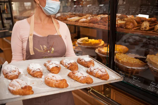 新鮮な焼き菓子のクロワッサンを持って 医療面マスクを身に着けている女性パン屋の作物のショット — ストック写真