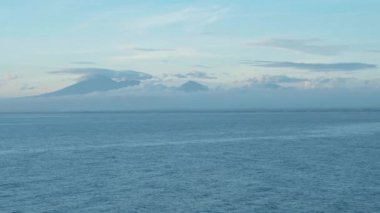 Mavi berrak su, bulutlar ve ufukta volkanlar olan deniz manzarasının videosu.