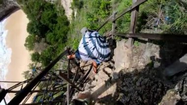 Eski bir kaya basamağındaki uygun bir insanın, yüksek bir dağdaki uçuruma deniz ve kaya manzaralı Kelingking plajındaki ilk kişi görüntüsü.