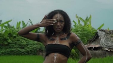 Güzel, esmer Afrikalı bir kızın videosu. Makyajlı ve vücuduna etnik çizgiler çizmiş. Tropikal bir bahçede yürüyor.