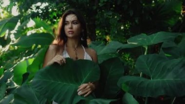 Güzel, genç, esmer Avrupalı bir kızın videosu. Makyajlı ve vücuduna etnik çizgiler çizmiş. Tropikal bir bahçede yürüyor.