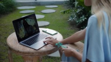 Tahta bir masada oturan ve yeşil bir bahçede dizüstü bilgisayarın arkasında çalışan genç bir kızın videosu.