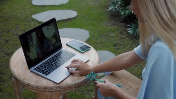木製のテーブルに座って緑の庭でノートパソコンの後ろで働いている若い女の子のビデオ — ストック動画