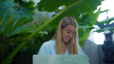 Tahta bir masada oturan ve yeşil bir bahçede dizüstü bilgisayarın arkasında çalışan genç bir kızın videosu.