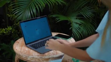 Tahta bir masada oturan ve yeşil bir bahçede dizüstü bilgisayarın arkasında çalışan genç bir kızın videosu.