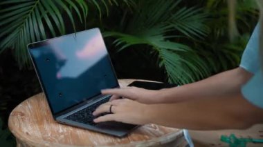 Tahta bir masada oturan ve yeşil bir bahçede dizüstü bilgisayarın arkasında çalışan genç bir kızın videosu.