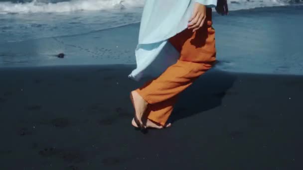 背景に海の波と黒砂に沿って自然の中で裸足で走る黄色のパンツの若い女の子 — ストック動画