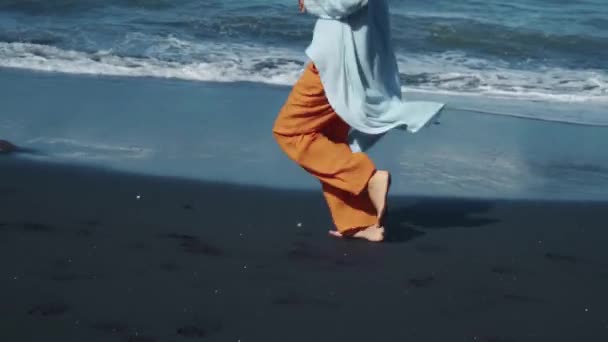 背景に海の波と黒砂に沿って自然の中で裸足で走る黄色のパンツの若い女の子 — ストック動画