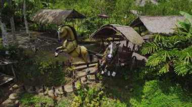 Yeşil bir vadiden çekilen insansız hava aracı videosu. Bali adasında, içinde at heykelleri olan palmiye ormanının ortasında duran bir ev.