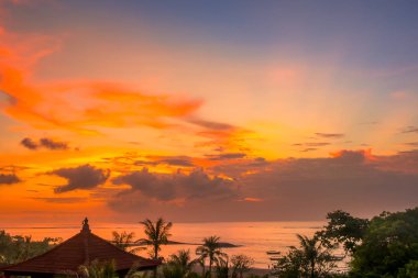 Endonezya. Bali Adası. Plaj ve okyanus üzerinde renkli gökyüzü günbatımı. Tekneler, palmiye ağaçları ve geleneksel çatı