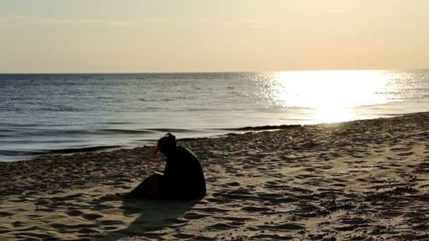 乌克兰 152017 空的海滩在日落 无法辨认的女孩坐在沙滩上看书 小波浪和太阳的踪影在水上 — 图库视频影像