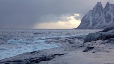 Kış Norveç. Kuzey Lofoten. Bulutlu akşam. Karla kaplı dağlar ve kayalık sahil. Sörf dalgası. Ağır çekim